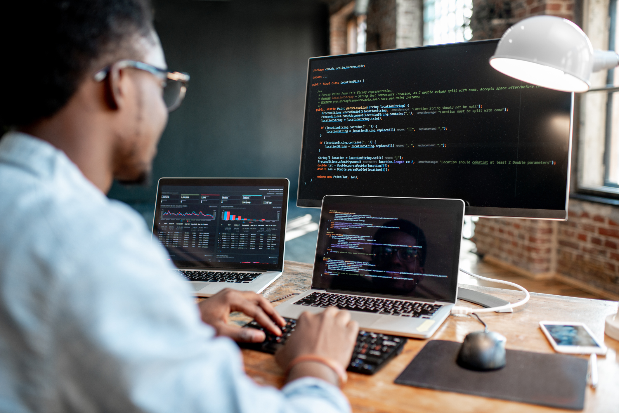 Young male programmer writing program code sitting at the workplace with three monitors in the office. Image focused on the screen