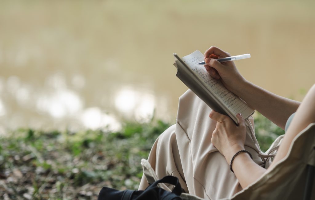 A woman journaling in her book as she reflects on her thoughts while sitting quietly in the forest and enjoying the nature
